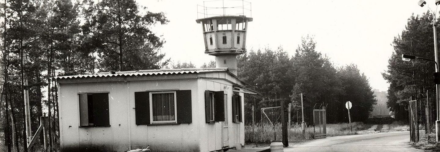 Das Bild zeigt ein kleines Wachhäuschen mit Wellblechdach vor einem Wachturm neben einer Straße. Diese führt durch ein geöffnetes Tor. Die Umgebung ist mit Nadelbäumen bewaldet. Vor dem Wachhäuschen stehen zwei Mülltonnen.