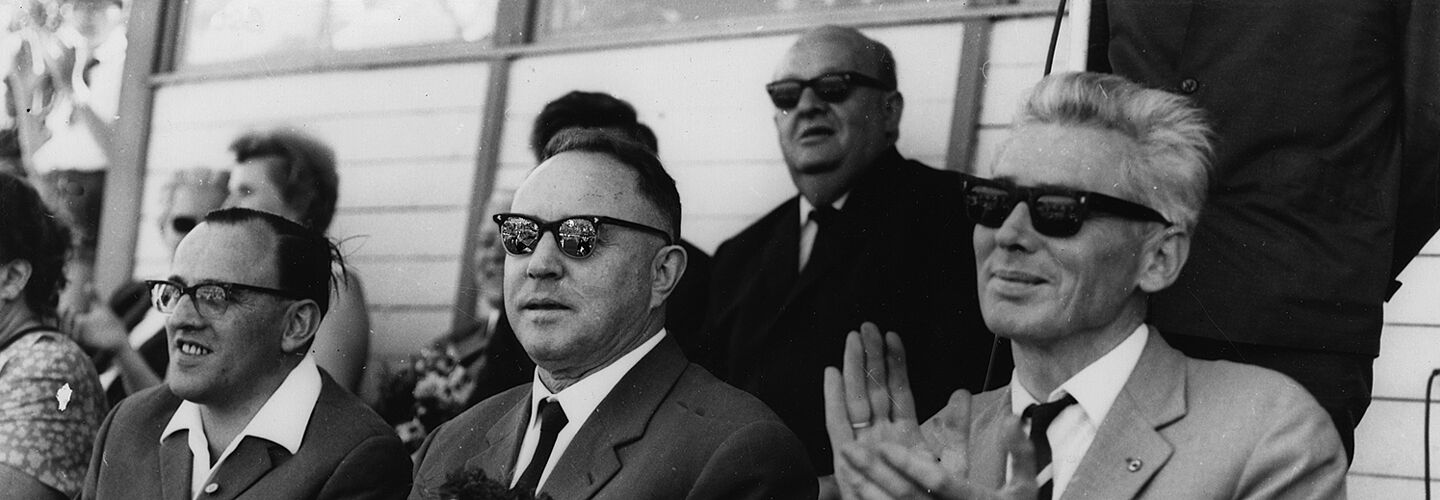 Schwarzweißfoto: Eine Reihe älterer Herren in Anzügen und mit Sonnenbrillen sitzen applaudierend auf einer Tribüne.