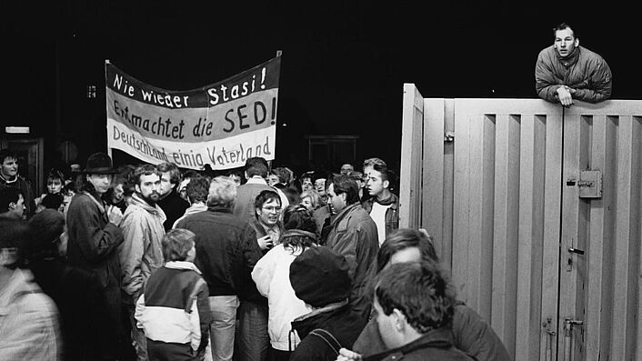 Das Schwarz-Weiß-Foto zeigt eine Gruppe von Menschen am geöffneten Haupteingang der Stasi-Zentrale an der Ruschestraße/Normannenstraße. Auf einem Banner steht 'Nie wieder Stasi! Entmachtet die SED! Deutschland einig Vaterland'. Oben rechts im Bild lehnt sich ein Mann über das Eingangstor.