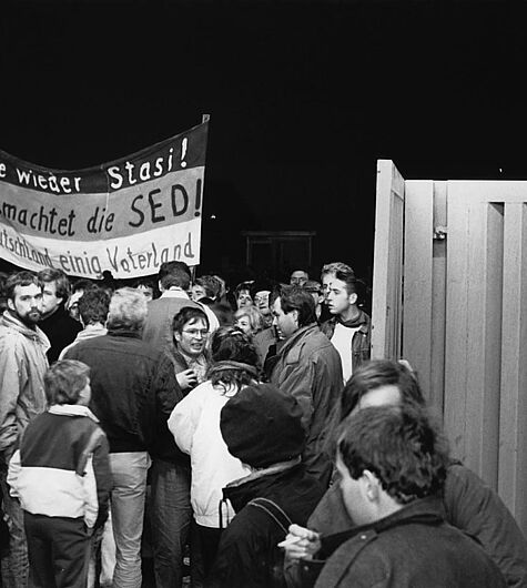 Das Schwarz-Weiß-Foto zeigt eine Gruppe von Menschen am geöffneten Haupteingang der Stasi-Zentrale an der Ruschestraße/Normannenstraße. Auf einem Banner steht "Nie wieder Stasi! Entmachtet die SED! Deutschland einig Vaterland". Oben rechts im Bild lehnt sich ein Mann über das Eingangstor.  , Quelle:
                    Foto: Andreas Schoelzel