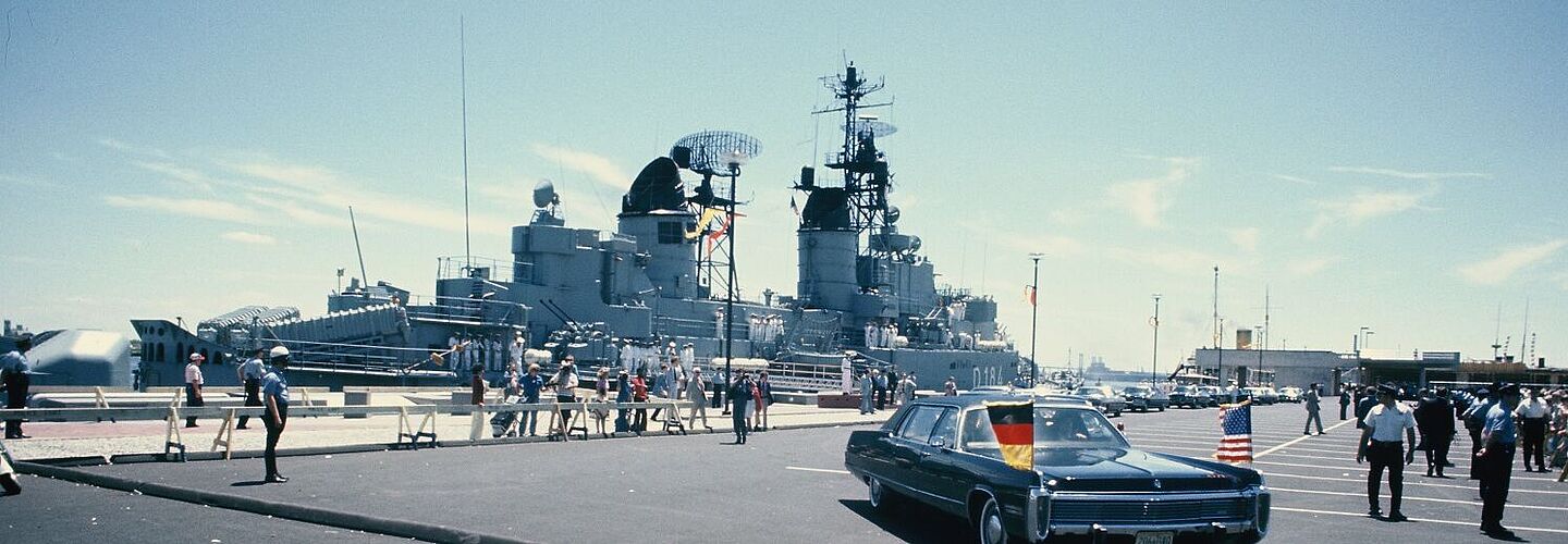 Ein großes Schiff liegt in einem Hafen vor Anker. Im Vordergrund ist ein Pkw mit deutscher und US-amerikanischer Flagge zu sehen.