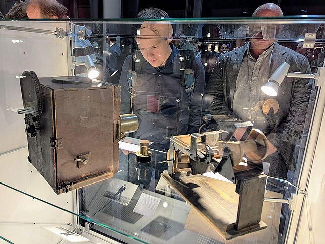 Zwei Personen sind der Kamera zugewandt und schauen in eine Vitrine, in der eine altertümliche Kamera und ein zweites Objekt mit Rollen zu sehen sind.