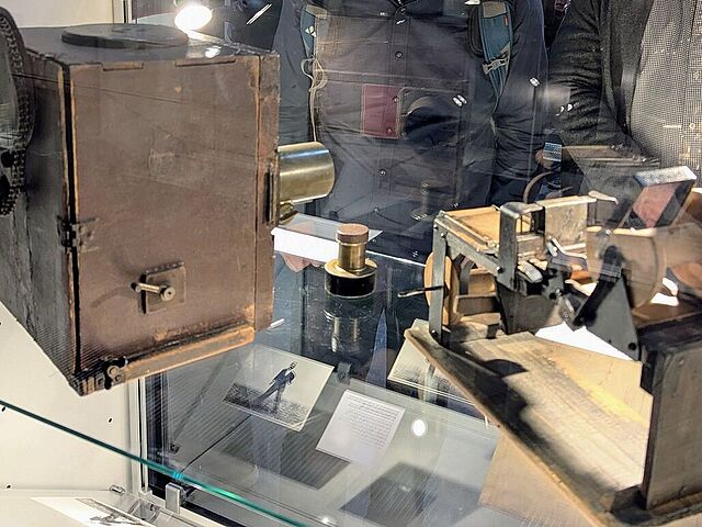 Zwei Personen sind der Kamera zugewandt und schauen in eine Vitrine, in der eine altertümliche Kamera und ein zweites Objekt mit Rollen zu sehen sind.