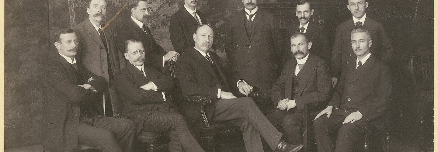 Das schwarz-weiße Bild zeigt elf Männer, aufgestellt in zwei Reihen. Die fünf Männer der vorderen Reihe sitzen auf Stühlen, der Rest steht hinter ihnen. Die Männer tragen Anzüge und haben große Schnurbärte. Der Präsident Anton Geißen sitzt in der Mitte.