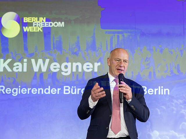 Ein Mann im Anzug spricht in ein Mikrofon. Hinter ihm ist eine beleuchtete Wand mit den Worten "Berlin Freedom Week Kai Wegner"