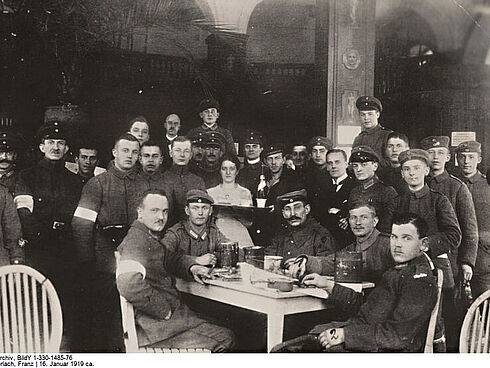 An einem Tisch sitzen fünf Männer, die zum Teil Uniformen, Mützen und Armbinden tragen und in die Kamera schauen. Um den Tisch herum stehen 23 Männer und blicken ebenfalls in die Richtung des Fotografen. Es handelt sich um eine Schwarz-Weiß-Aufnahme vom Januar 1919.