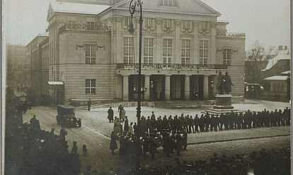 Schwarz-Weiß-Aufnahme einer Menschenschlange entlang der Vorderfront eines historischen Gebäudes