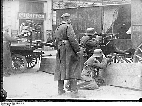 Mehrere Männer mit langen Mänteln und Helmen stehen und hocken mit Gewehren im Anschlag hinter einer Kutsche und einem dicken Holzbrett, die als Barrikade dienen. Es handelt sich um eine Schwarz-Weiß-Aufnahme vom Januar 1919.