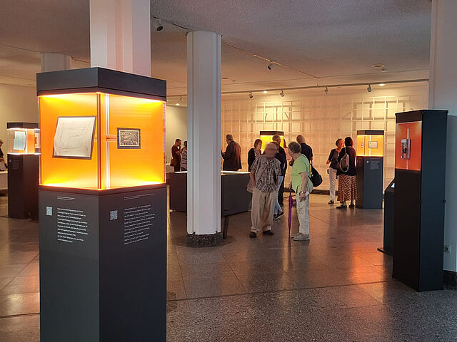 Mehrere Personen in einen halbdunklen Raum mit orange beleuchteten Ausstellungsstelen.