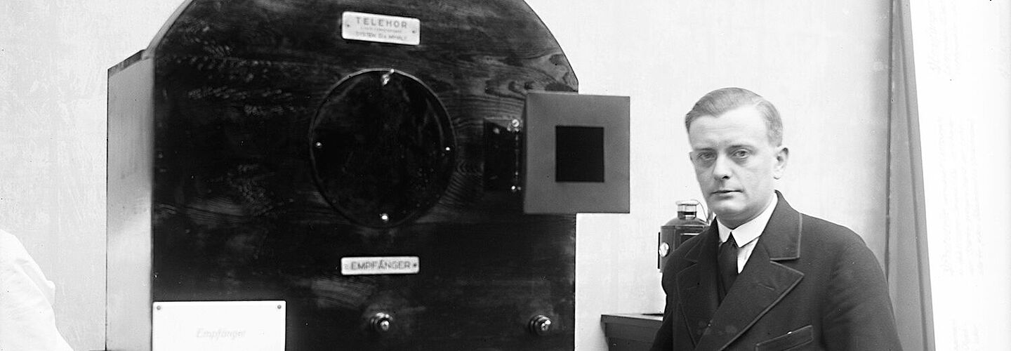 Schwarz-Weiß-Aufnahme mit einem Mannes im Anzug neben einem antiken Fernsehapparat