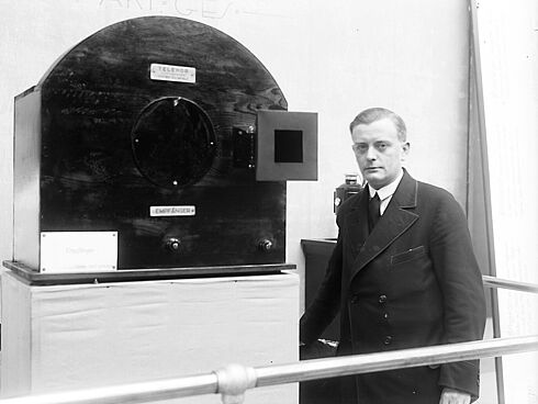 Schwarz-Weiß-Aufnahme mit einem Mannes im Anzug neben einem antiken Fernsehapparat