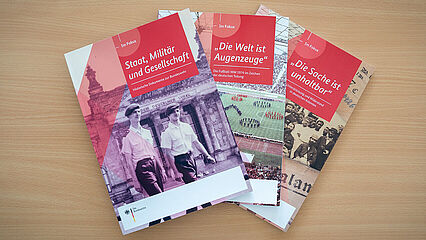 Drei Hefte liegen übereinander auf einem Tisch. Auf dem Cover des obersten Hefts sind zwei Bundeswehrsoldaten vor dem Reichstagsgebäude in Berlin zu sehen. Auf dem Cover darunter ist ein Fußballstadion und auf dem Cover des untersten Hefts sind mehrere Menschen abgebildet.