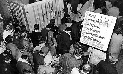 Das Bild zeigt Menschen, die durch ein geöffnetes Tor strömen und Plakate bei sich tragen. Auf einem ist zu lesen: Stasi / Einheits / Diktatoren / Probieren / Demokratischen / Sozialismus / Nie Wieder '