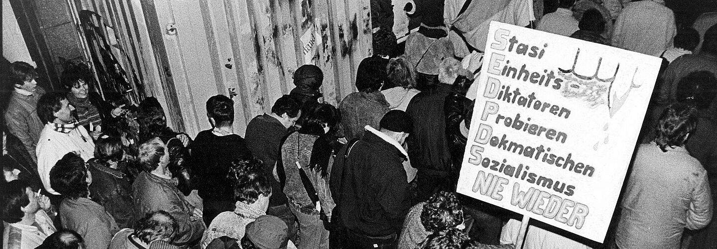 Das Bild zeigt Menschen, die durch ein geöffnetes Tor strömen und Plakate bei sich tragen. Auf einem ist zu lesen: Stasi / Einheits / Diktatoren / Probieren / Demokratischen / Sozialismus / Nie Wieder '