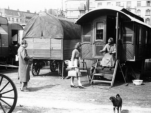 Schwarz-Weiß-Aufnahme eines Mannes und einer Dame, die mit einer älteren Frau, die auf einer Holztreppe eines Wohnwagens sitzt, sprechen