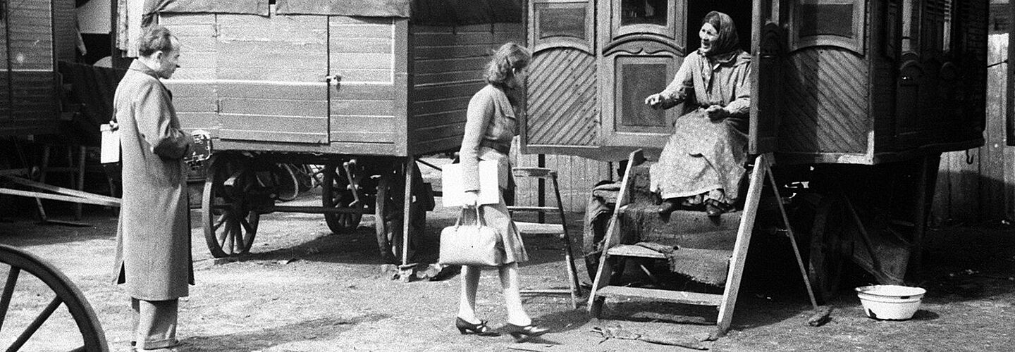 Schwarz-Weiß-Aufnahme eines Mannes und einer Dame, die mit einer älteren Frau, die auf einer Holztreppe eines Wohnwagens sitzt, sprechen