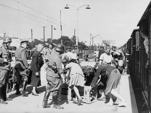 Polnische Zivilistinnen und Zivilisten be- oder entladen eine Kiste vor einem Zug mit Güterwagen am Bahnhof Włochy. Daneben stehen deutsche Soldaten.