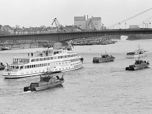 Schwarz-Weiß-Aufnahme eines großen Fahrgastschiffes, das in Begleitung kleinerer militärischer Boote auf dem Rhein unter einer Brücke hindurchfährt.
