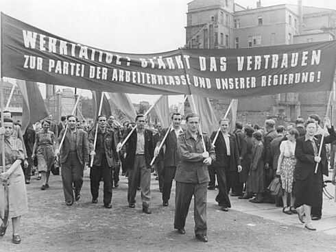 Das Bild zeigt demonstrierende Menschen. Sie tragen ein Banner mit der Aufschrift "Werktätige: Stärkt das Vertrauen zur Partei der Arbeiterklasse und unserer Regierung"