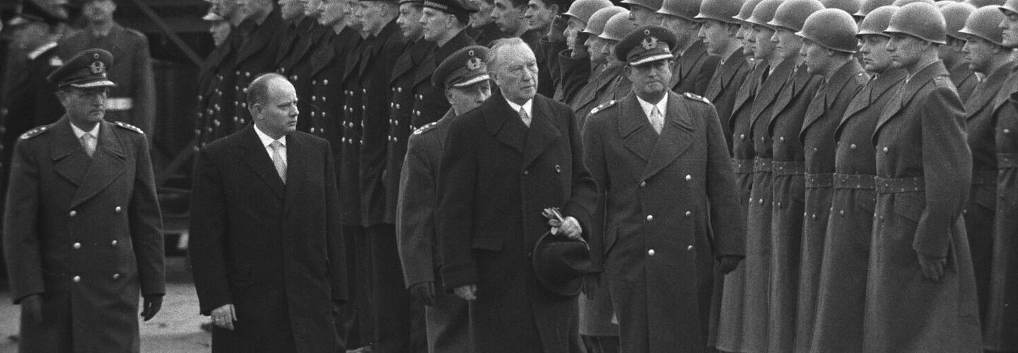 Bundeskanzler Konrad Adenauer und Verteidigungsminister Theodor Blank schreiten bei ihrem Besuch in Andernach zusammen mit drei hochrangigen Bundeswehroffiziere eine angetretene Formation von Soldaten des Heeres und der Marine ab. Im Hintergrund stehen Pressefotografen.