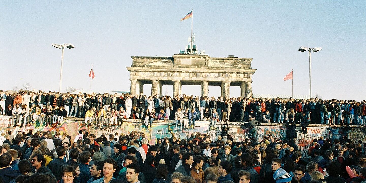 Menschen aus West- und Ostberlin auf der Mauerkrone am Brandenburger Tor, 10.11.1989 Bild mit Menschen aus West- und Ostberlin auf der Mauer am Brandenburger Tor, 10.11.1989 , Quelle:
Bundesregierung, B 145 Bild-00100901 / Lehnartz, Klaus