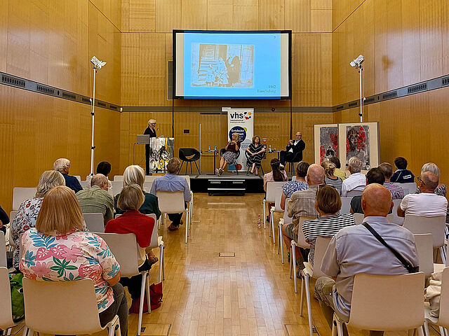 Blick in einen Saal mit Spitzdach - im Hinertgrund eine Bühne mit vier Personen, im Vordergrund sitzt das vom Betrachtenden abgewandte Publikum