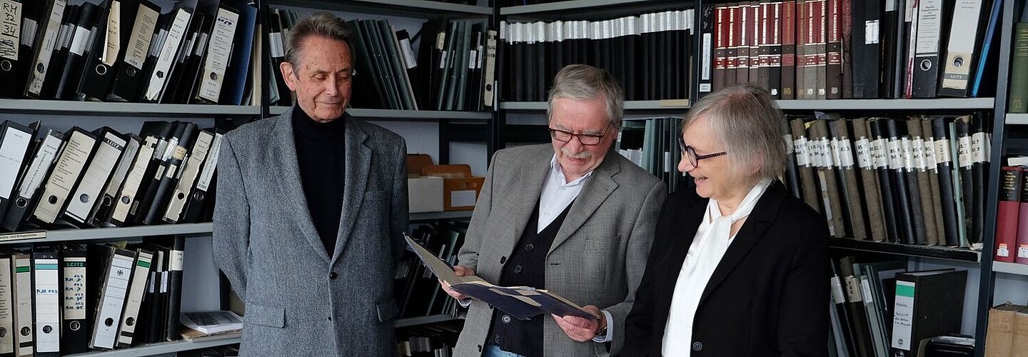 Zwei Männer und eine Frau stehen nebeneinander an einem Tisch mit zahlreichen Akten. Der Mann in der Mitte hält eine aufgeschlagene Akte in der Hand, in die alle drei Personen hineinschauen.