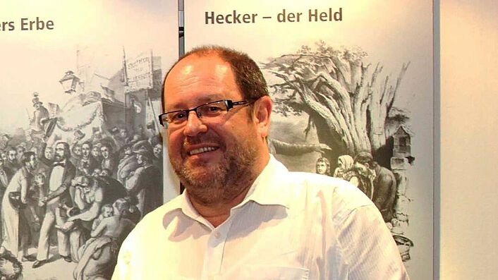 Ein Mann mit Brille steht in einem weißen Hemd vor einem Roll-Up und lächelt. Er hält ein kleines Buch in die Kamera.