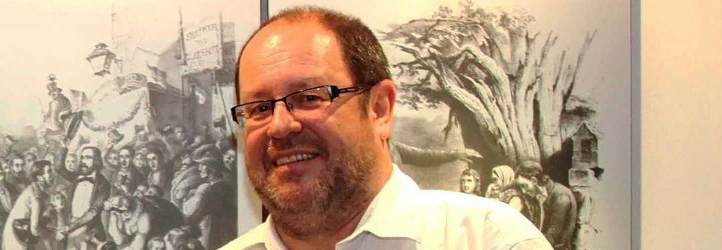 Ein Mann mit Brille steht in einem weißen Hemd vor einem Roll-Up und lächelt. Er hält ein kleines Buch in die Kamera.