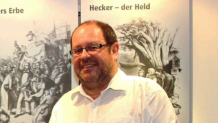 Ein Mann mit Brille steht in einem weißen Hemd vor einem Roll-Up und lächelt. Er hält ein kleines Buch in die Kamera.