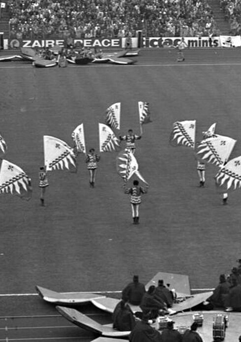 Eröffnungsfeier Auf dem Spielfeld stehen mehrere Frauen mit kurzen Kleidern, die je zwei große Fahnen schwingen. An den langen Spielfeldrändern sitzen Musiker mit ihren Instrumenten.