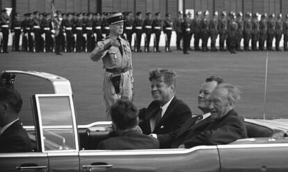 John F. Kennedy, Willy Brandt und Konrad Adenauer sitzen lachend in einem Auto. Im Hintergrund sind mehrere Soldaten zu sehen.