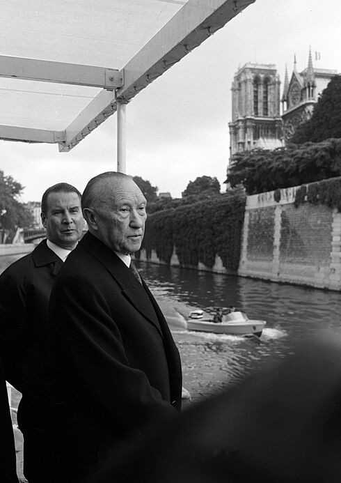 Adenauer und Schröder stehen an der Reling eines Ausflugsschiffs und schauen sich die Umgebung an. Im Hintergrund ist ein schlossähnliches Bauwerk zu sehen, auf dem Wasser ein kleines Boot.