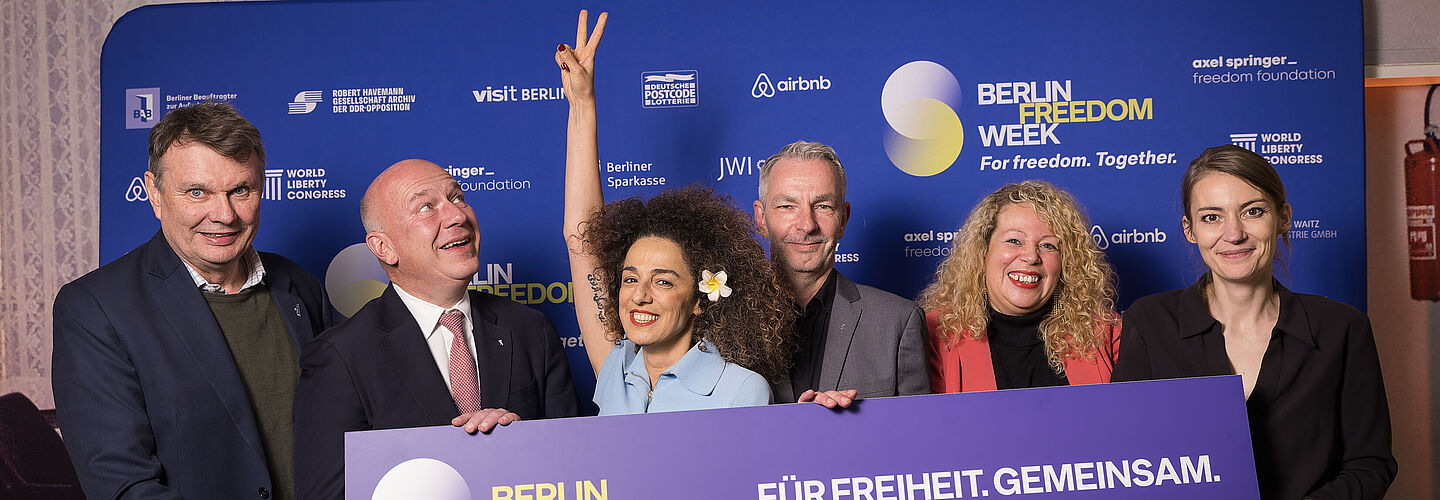 Eine Gruppe von Personen steht vor einem blauen Hintergrund und hält ein Schild hoch mit der Aufschrift 'Berlin Freedom Week Für Freiheit. Gemeinsam. 8.-15. November 2025
