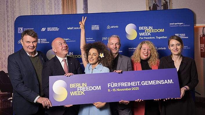Eine Gruppe von Personen steht vor einem blauen Hintergrund und hält ein Schild hoch mit der Aufschrift 'Berlin Freedom Week Für Freiheit. Gemeinsam. 8.-15. November 2025