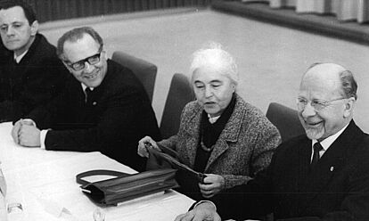 Schwarz-Weiß-Fotografie mit drei Herren und einer Frau in formeller Kleidung, lächelnd und an einem langen Tisch sitzend