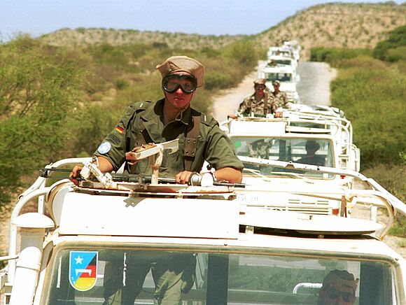 Ein Bundeswehrsoldat mit Wüstenbrille und Tropenhut blickt aus der Dachluke eines Bundeswehrfahrzeugs. Dieses bildet mit einer Reihe von anderen Fahrzeugen im Hintergrund einen Konvoi.