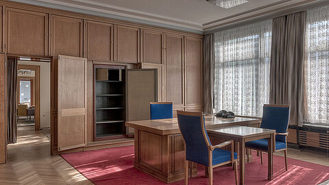 Das Bild zeigt ein hochwertig eingerichtetes Büro mit Holzverkleidung. In der Mitte des Raumes steht ein großer Schreibtsich mit drei Stühlen. Die Fenster sind mit halbdurchlässigen Vorhängen abgedeckt