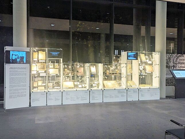 Mehrere Vitrinen vor einer Glasfront sind zu sehen. Sie sind mit verschiedenen Kameras und Dokumenten gefüllt.