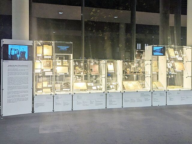Mehrere Vitrinen vor einer Glasfront sind zu sehen. Sie sind mit verschiedenen Kameras und Dokumenten gefüllt.