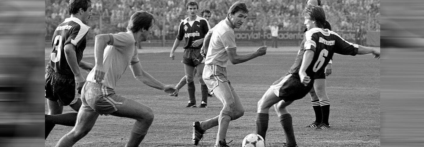 Schwarz-weiß Foto: fünf Fußballspieler, die um einen Ball dribbeln, im Hintergrund gefüllte Zuschauerränge.