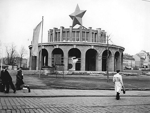 Im Hintergrund sieht man ein großes, rundes Gebäude mit Arkadenbögen. Auf dem Dach ist ein großer Stern befestigt. Im Vordergrund sieht man Menschen, die über eine Straße gehen.