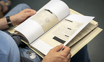 Ein Mann hat auf seinem Schoß eine Akte aufgeschlagen. Es lassen sich Schreibmaschinenschrift und ein Briefumschlag erkennen.