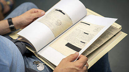 Ein Mann hat auf seinem Schoß eine Akte aufgeschlagen. Es lassen sich Schreibmaschinenschrift und ein Briefumschlag erkennen.