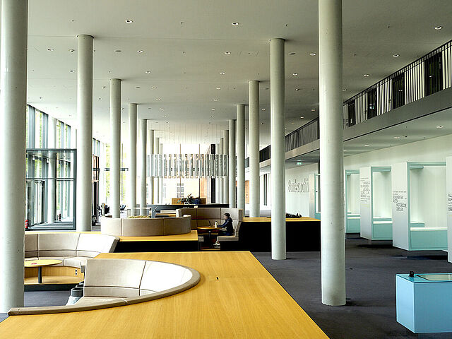 Blick in einen Raum mit zwei Reihen glatter Betonsäulen. Links eine Glasfront mit Tür, in der Mitte Sitzbereiche und im Hintergrund ein Tresen, rechts Ausstellungsvitrinen.