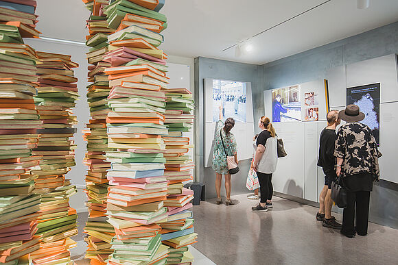 Vier Personen schauen sich die Bilder und Karten der Archivausstellung   "Einblick ins Geheime" an. In der Raummitte befindet sich ein Kunstwerk aus mehreren nachgebildeten Aktenstapeln. 