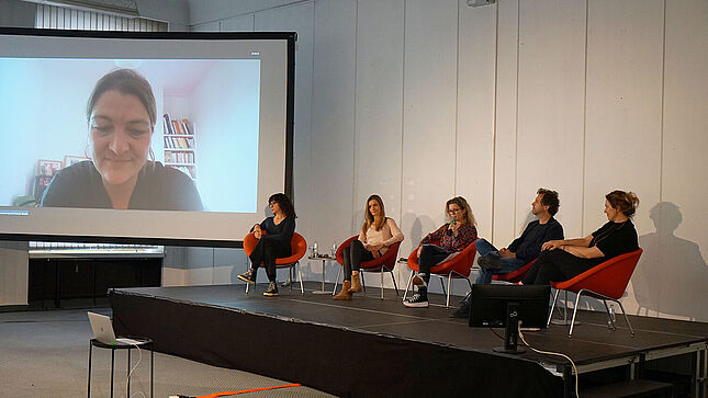 Die Podiumsgäste Nancy Biniadaki, Alexandra Finder, Nicolai Tegeler und Nadja Klier sowie Moderatorin Dagmar Hovestädt sitzen auf einer Bühne. Links neben der Bühne ist eine große Leinwand zu erkennen, auf der Luise Schröder zugeschaltet ist