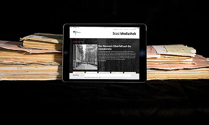 Das Bild zeigt die auf einem Tablet geöffnete Startseite der Stasi-Mediathek. Das Tablet lehnt an einigen Aken.