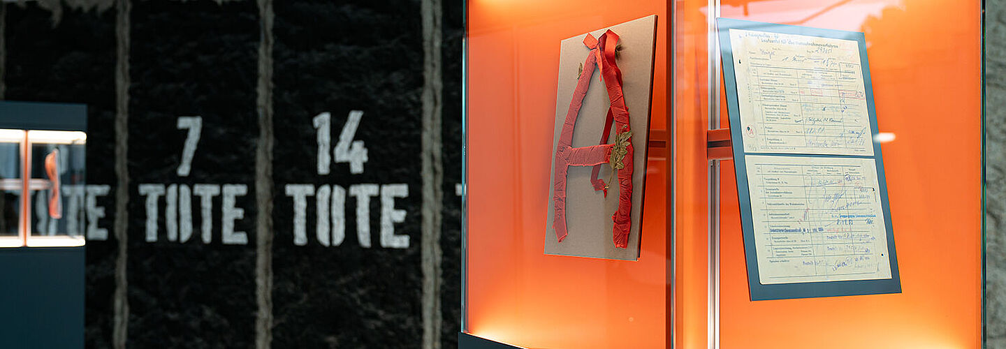 Eine Glasvitrine, in der sich ein aus Alufolie und Krepppapier gebasteltes „A“ sowie ein Dokument befinden.