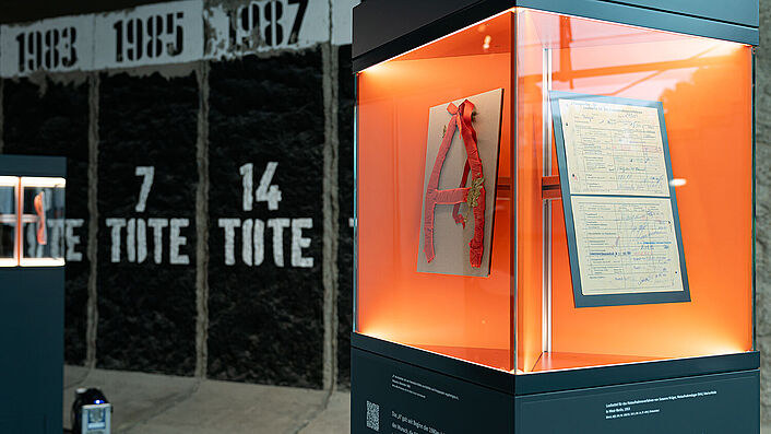 Eine Glasvitrine, in der sich ein aus Alufolie und Krepppapier gebasteltes „A“ sowie ein Dokument befinden.